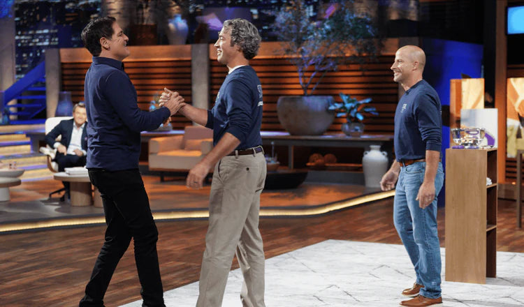 Two men shaking hands on a television show set with a third man standing nearby.