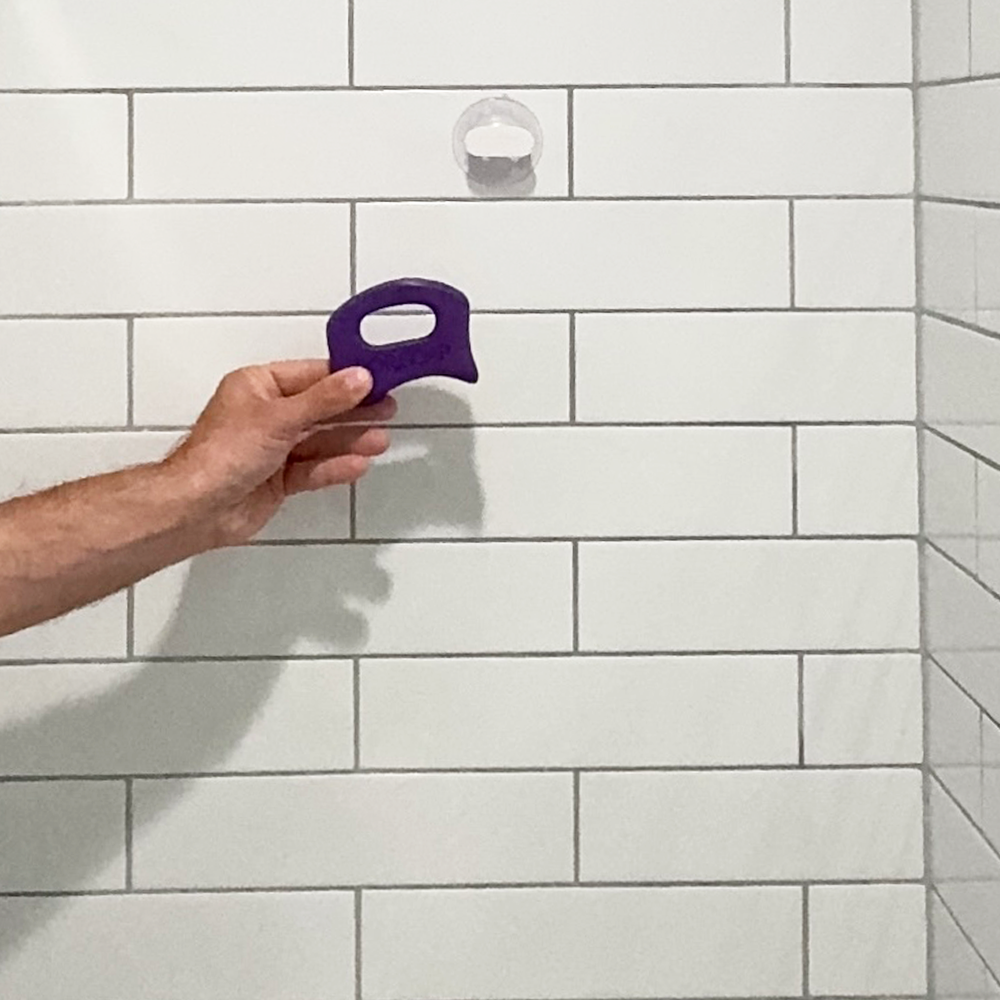Purple SORSOAP being placed on its suction cup holder, mounted on a clean tiled shower wall for easy access.
